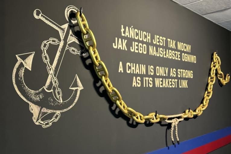 Dekoracja ściany: złoty łańcuch, kotwica i napis 'Łańcuch jest tak mocny jak jego najsłabsze ogniwo' w biurze. Ciemne tło, kreatywny design.