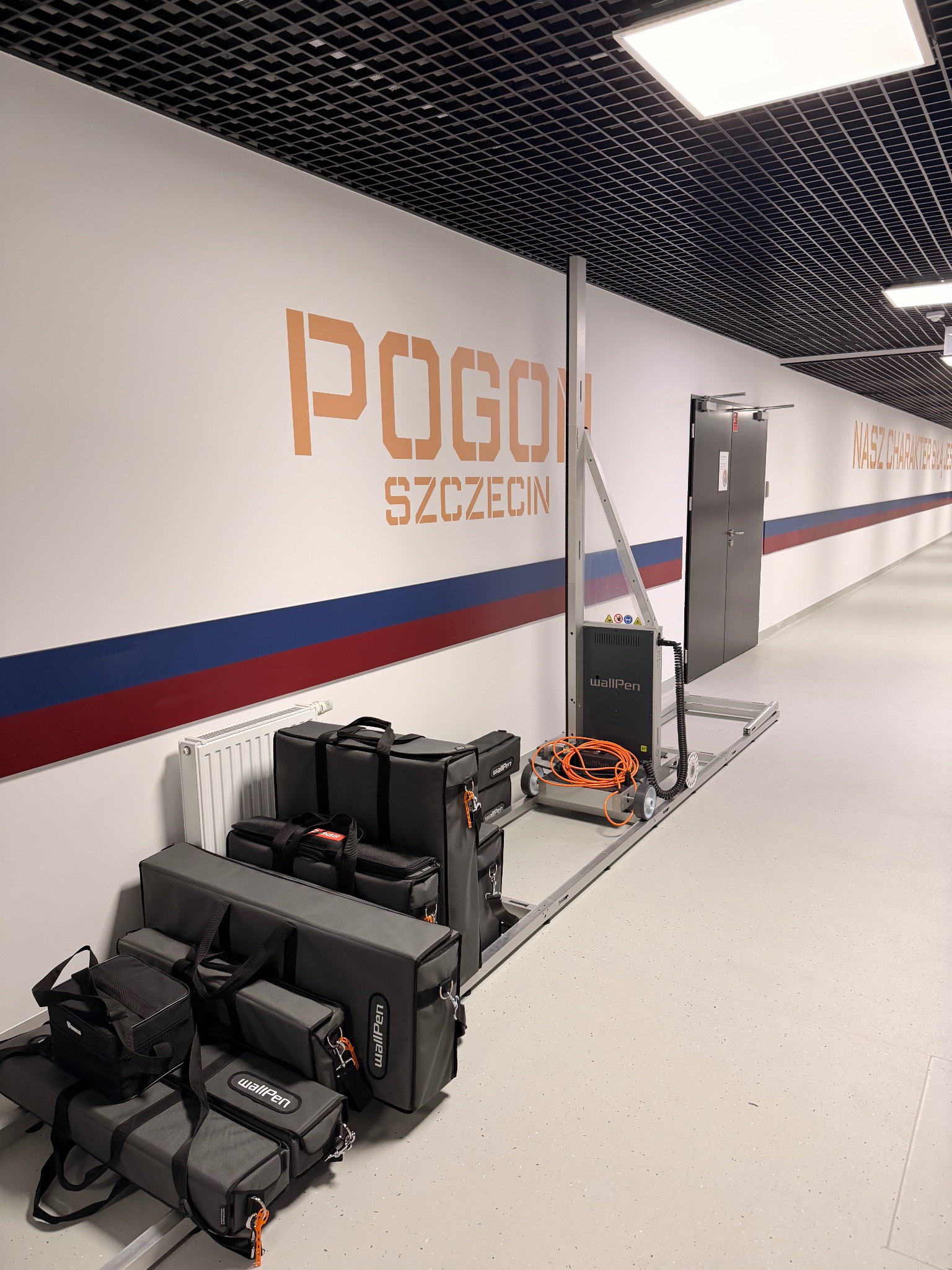 Wnętrze biura z napisem 'Pogoń Szczecin' malowanym na ścianie za pomocą urządzenia WallPen. Widoczne torby transportowe i sprzęt do malowania.