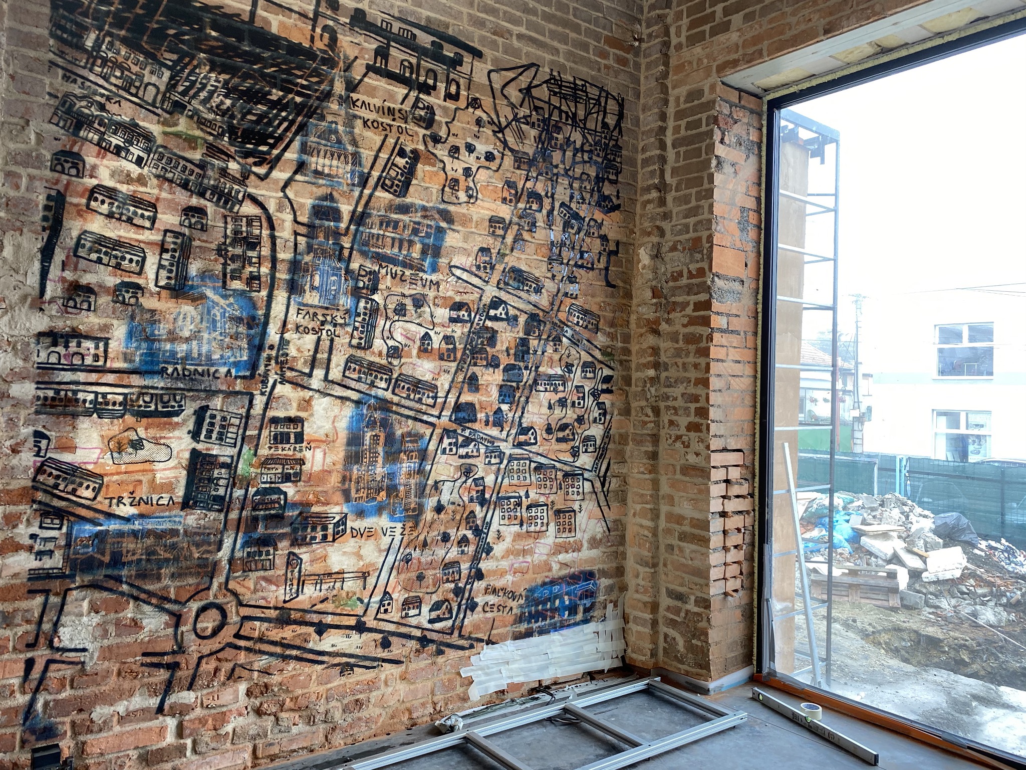 Mur z mapą miasta w stylu graffiti, widoczny przez otwarte okno w surowym wnętrzu. Elementy rusztowania i taśmy malarskiej sugerują prace wykończeniowe.