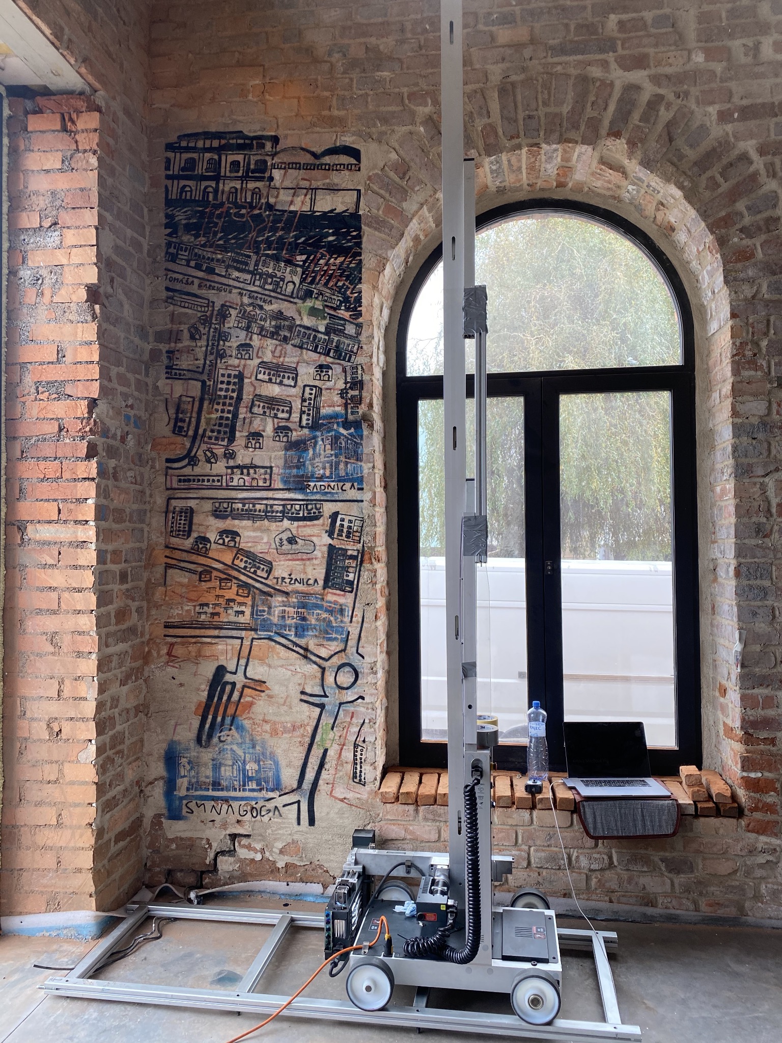 Automat do malowania ścian z naniesionym szkicem miasta na ceglanej ścianie w surowym wnętrzu. Laptop i woda na parapecie okna.