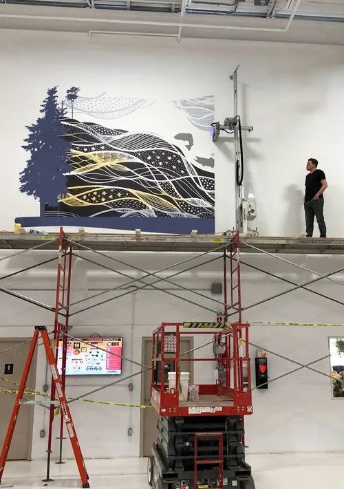 Malowanie artystyczne ściany w biurze za pomocą specjalistycznego robota. Na rusztowaniu stoi mężczyzna obserwujący proces. W tle widoczny ekran z informacjami.