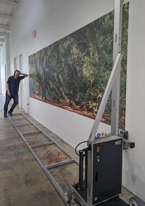 Wnętrze z maszyną do druku wielkoformatowego na ścianie, prezentującą leśny motyw. Mężczyzna opiera się o ścianę, obserwując proces drukowania.