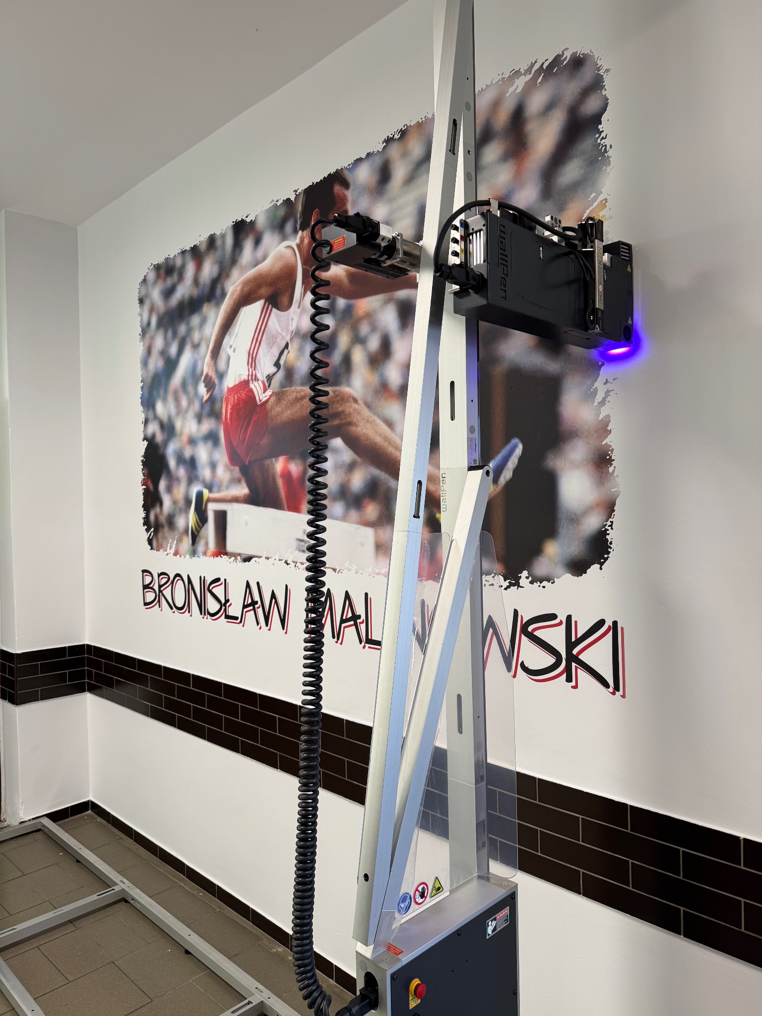 Drukarka Wallpen nanosi grafikę z Bronisławem Malinowskim na ścianę w Goleniowie. Nowoczesna technologia druku pionowego, detale wykończenia.