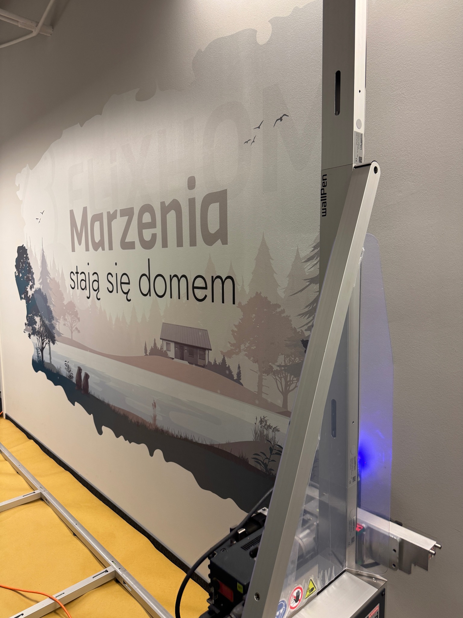 Ściana z nadrukiem krajobrazu wykonanym przez urządzenie WallPen. Napis: Marzenia stają się domem. Widoczne elementy urządzenia drukującego.