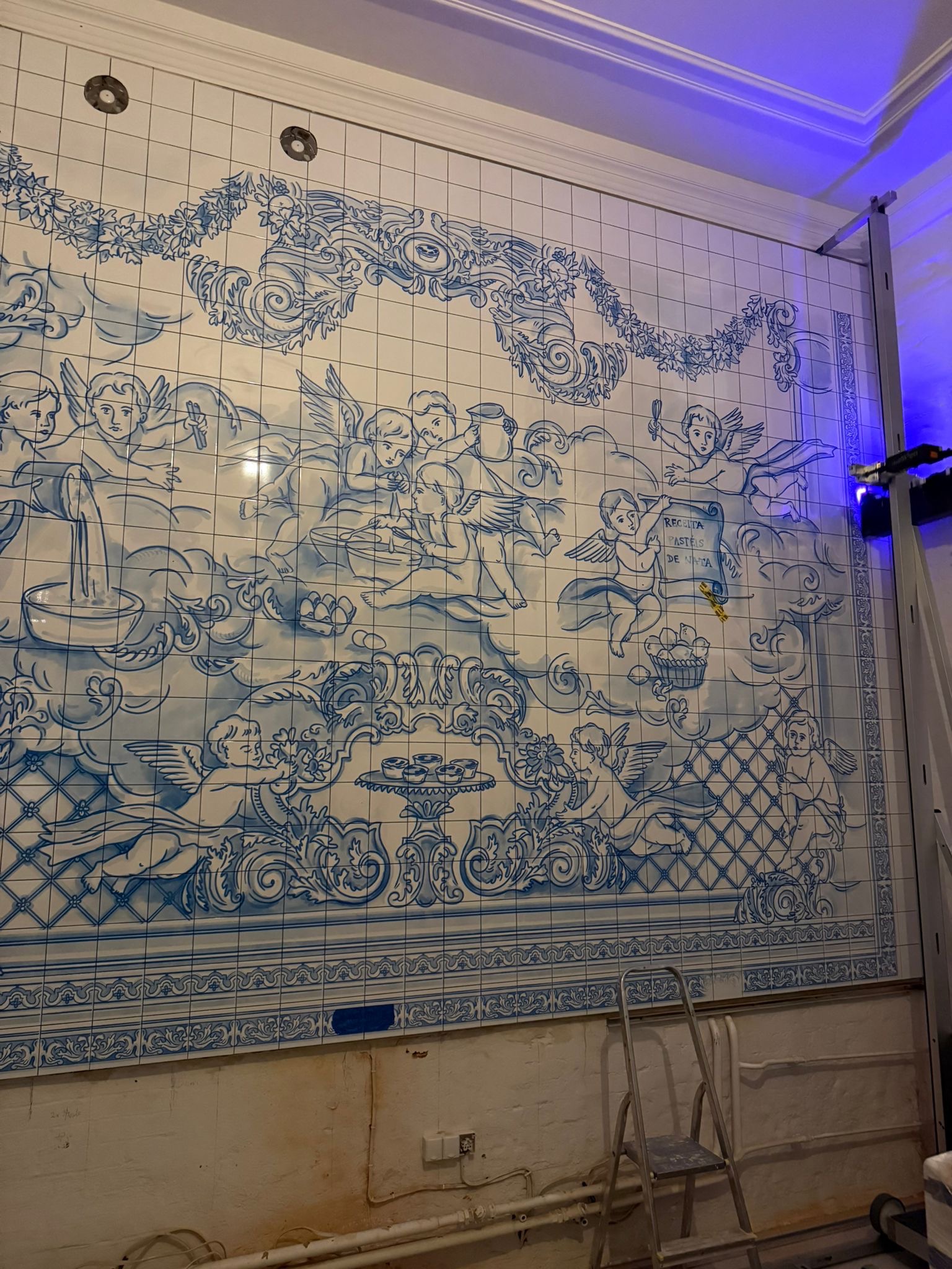 Ściana wyłożona białymi płytkami z niebieskim, ręcznie malowanym wzorem aniołów i ornamentów, nawiązującym do tradycyjnych portugalskich azulejos. Widoczna drabina i fragment rusztowania.