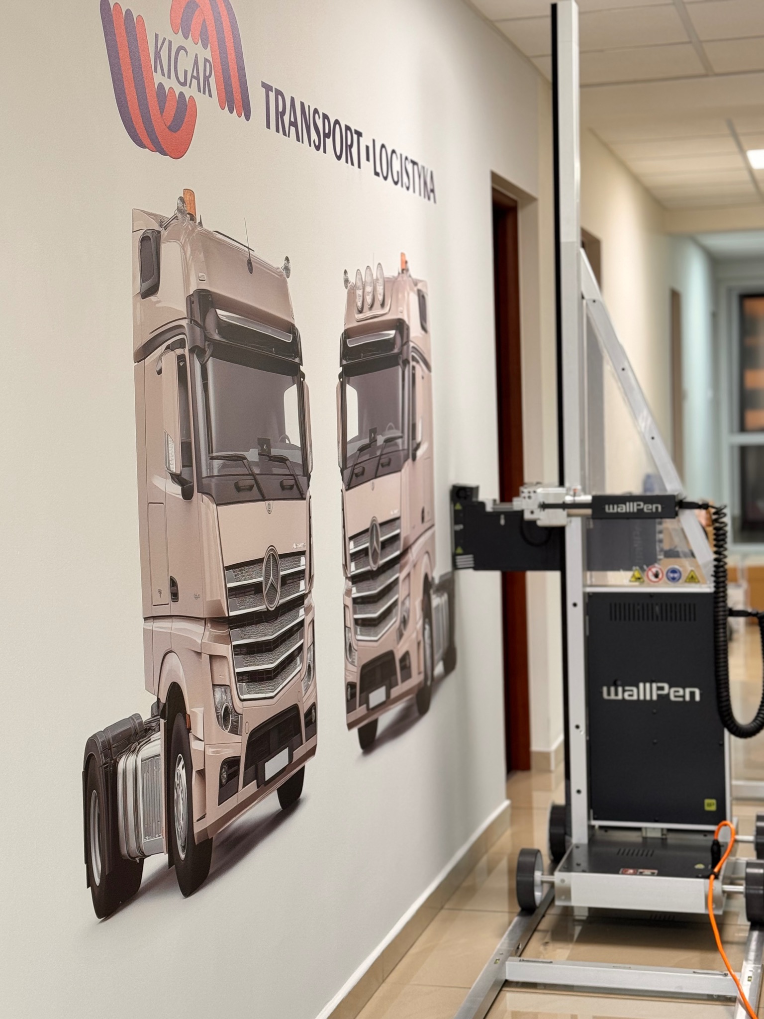 WallPen drukuje na ścianie w biurze grafiki dwóch ciężarówek Kigar Transport Logistyka. Drukarka na szynach, w tle drzwi i korytarz.