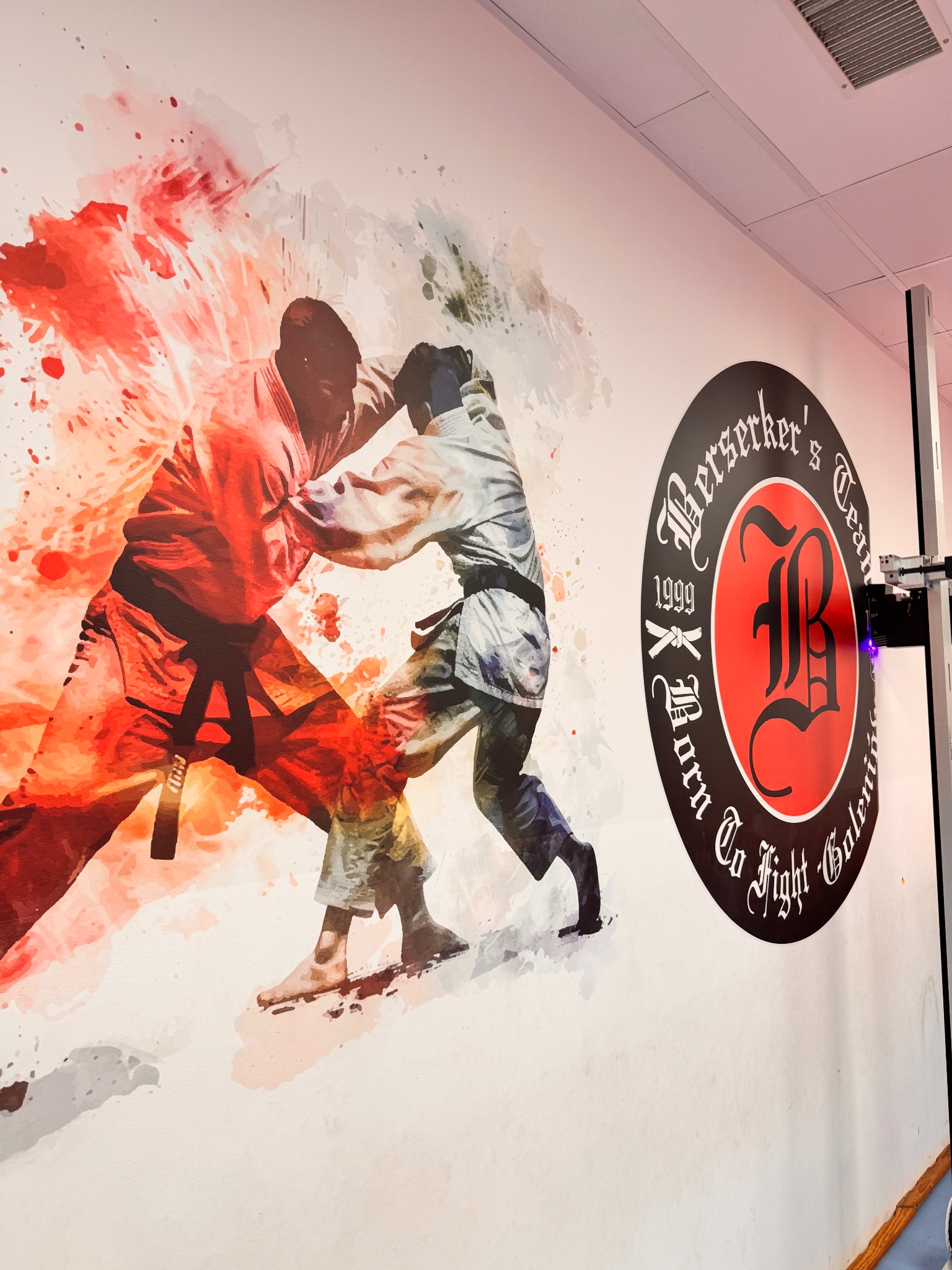 Malowidło ścienne w sali treningowej: dynamiczna ilustracja dwóch zawodników sztuk walki w kimonach, obok logo klubu 'Berserker's Team' z rokiem założenia 1999.