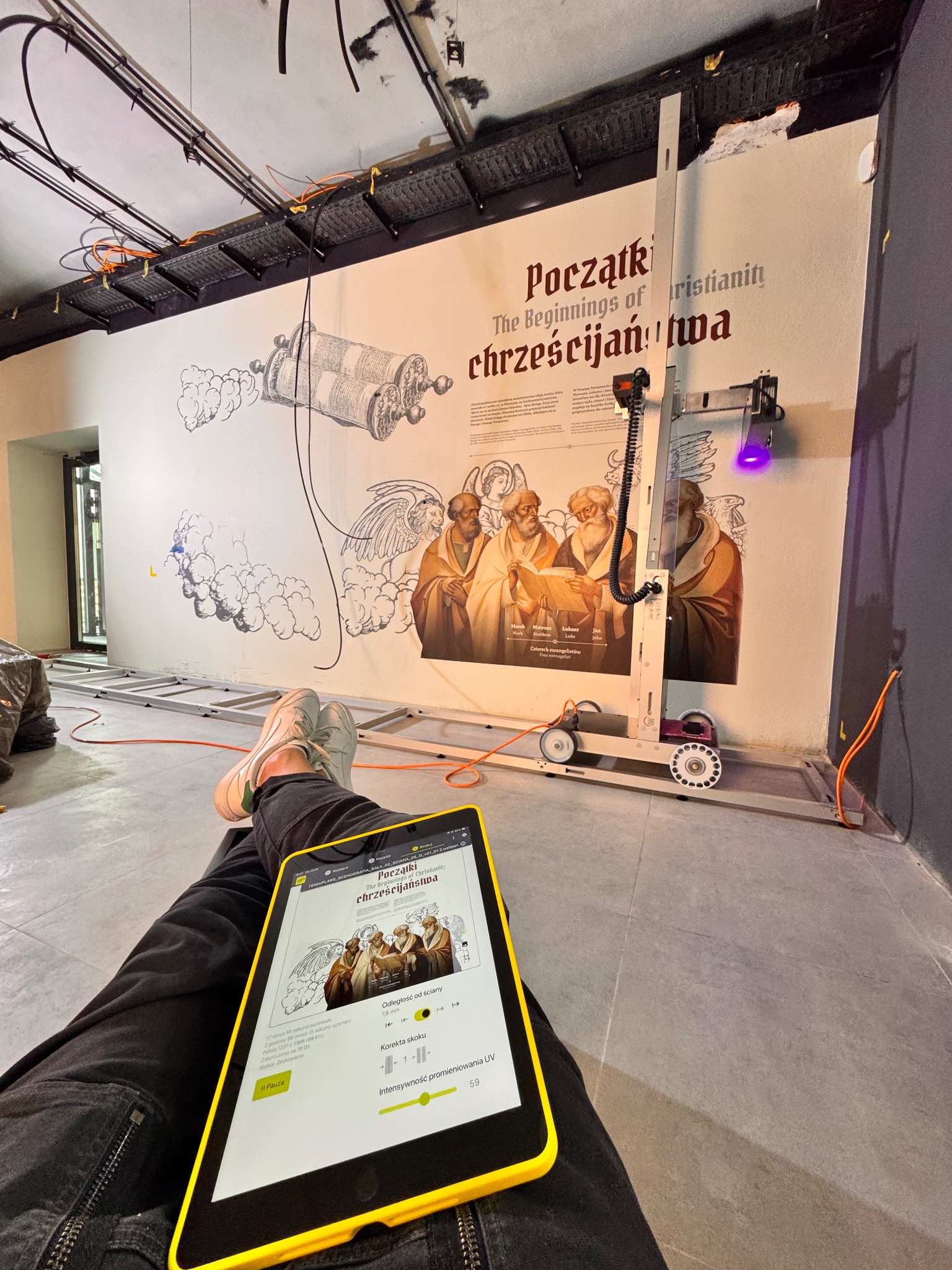Aplikacja na tablecie prezentuje grafikę ścienną 'Początki chrześcijaństwa' z użyciem mobilnego urządzenia UV do utwardzania farb. Widoczne instalacje pod sufitem.