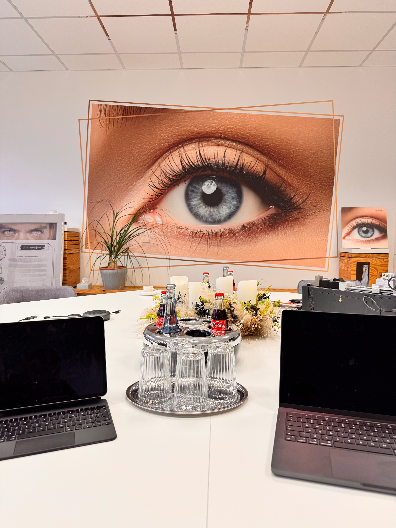 Wnętrze biura z plakatem oka na ścianie, dekoracjami na stole i dwoma laptopami. Nowoczesny design, jasne kolory, minimalistyczny styl. Elementy graficzne w tle.