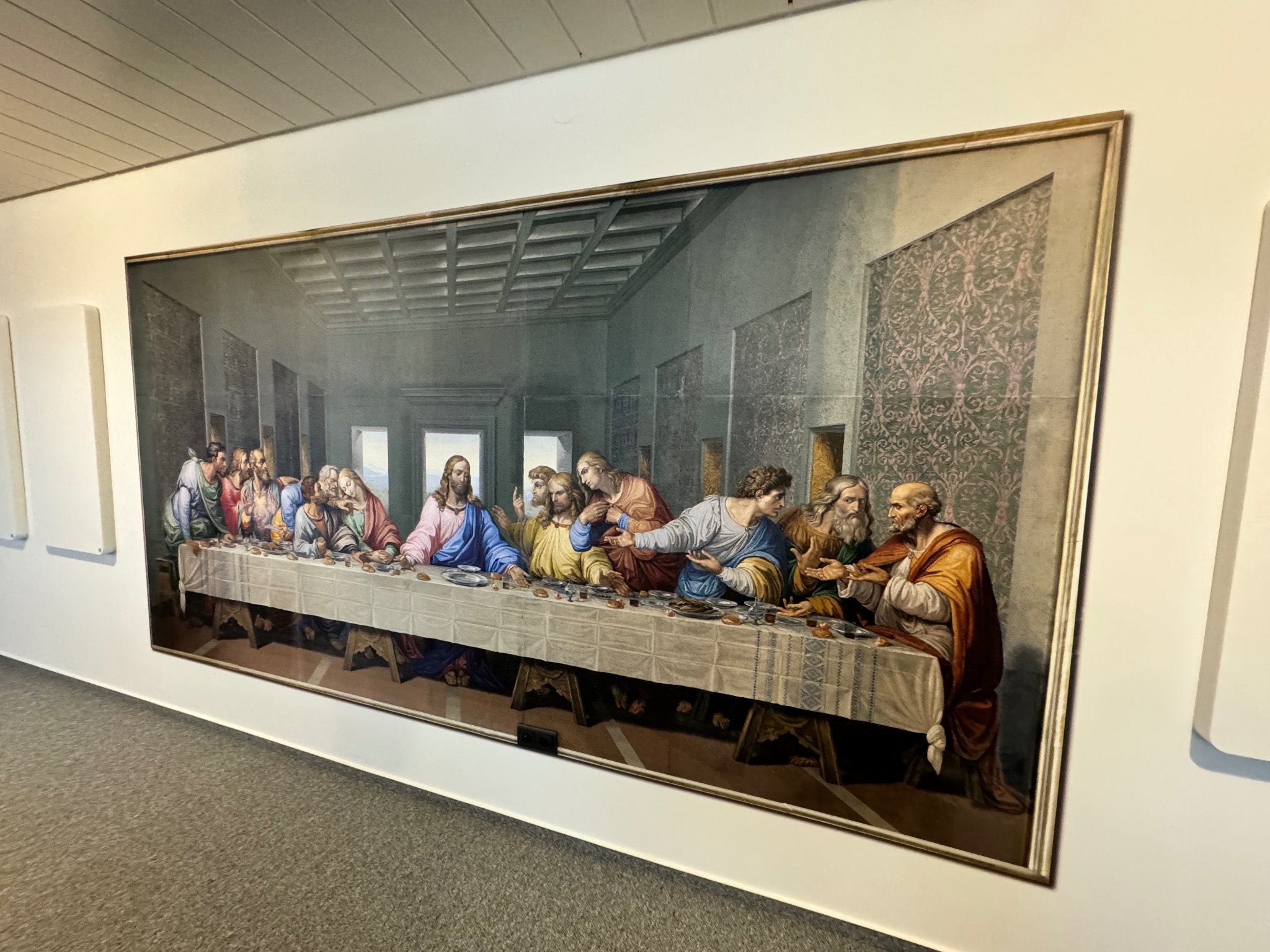 Reprodukcja Ostatniej Wieczerzy Leonarda da Vinci w złotej ramie, wisząca na białej ścianie w korytarzu. Widoczne elementy dekoracyjne i oświetlenie.