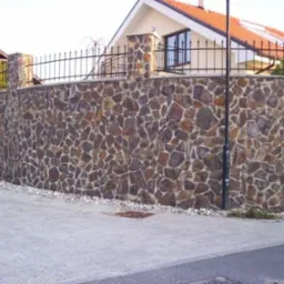 Masywny, łukowaty mur z kamienia elewacyjnego Andezyt, zwieńczony czarnym, kutym ogrodzeniem, chroniący posesję z widocznym fragmentem domu z czerwonym dachem.