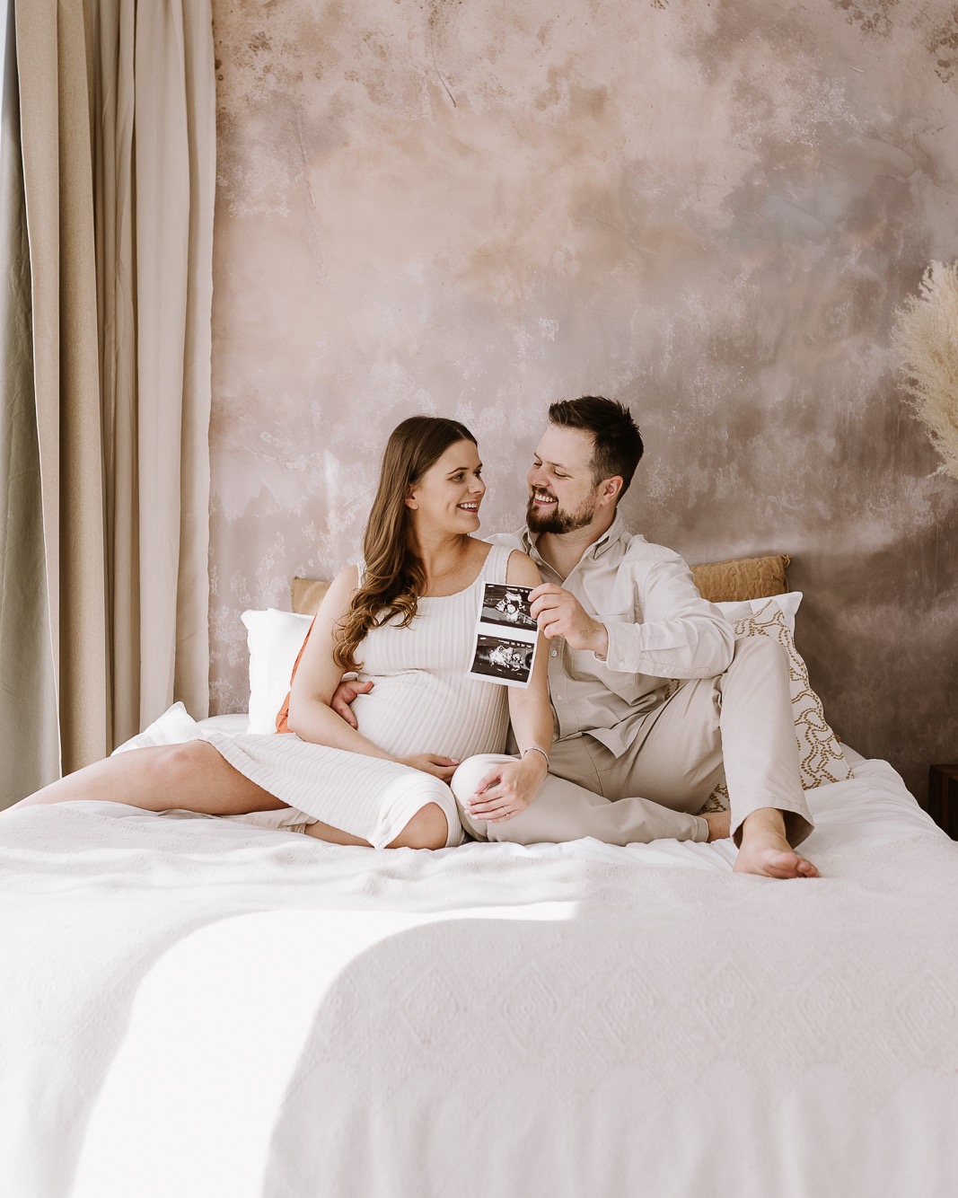 Uśmiechnięta para w jasnych ubraniach siedzi na łóżku, trzymając zdjęcie USG. Kobieta w ciąży, ciepłe światło, naturalne tło. Sesja w domowym klimacie.