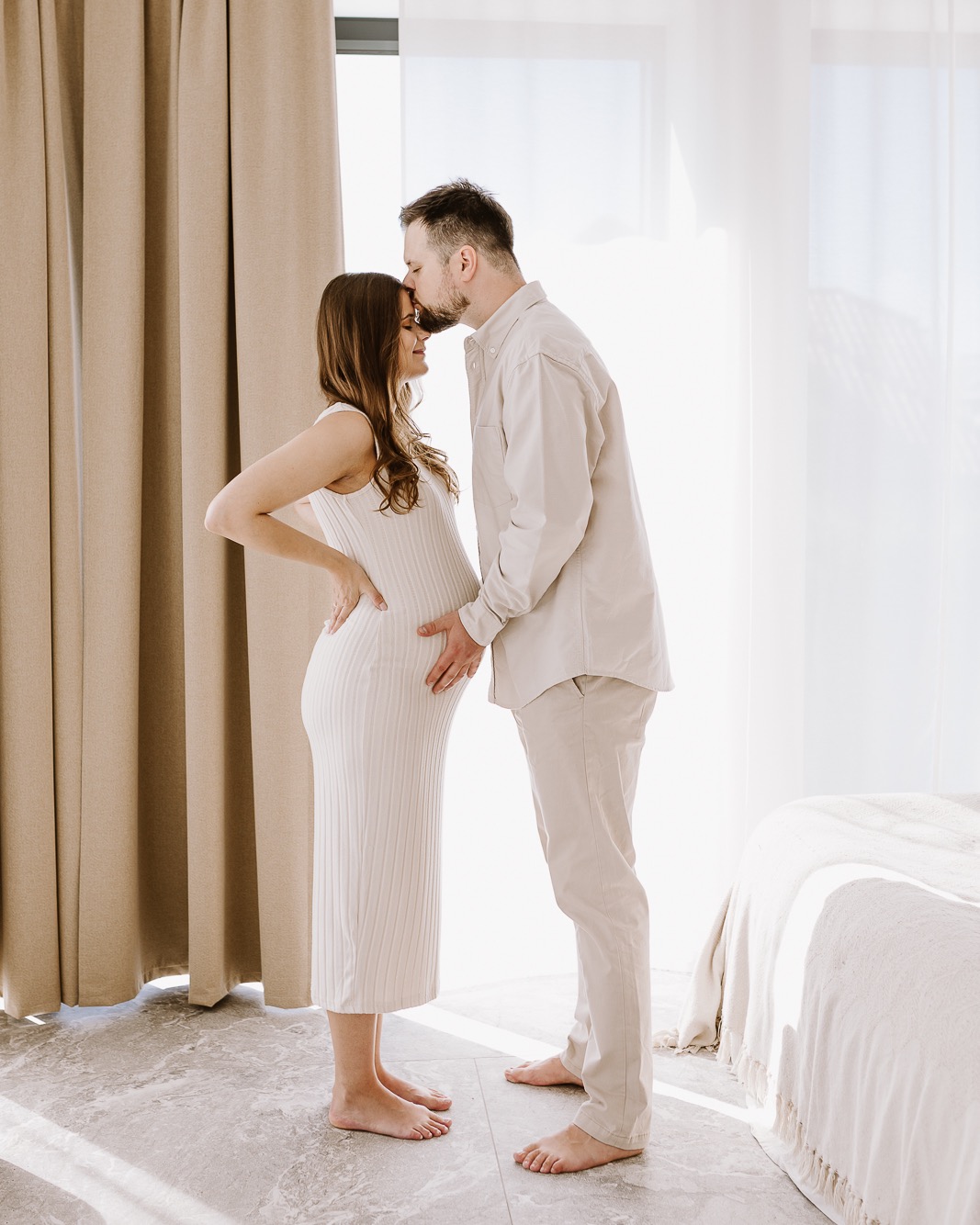 Czułe ujęcie: mężczyzna całuje w czoło kobietę w ciąży, oboje w jasnych ubraniach, dłoń mężczyzny na brzuchu kobiety, stonowane wnętrze z zasłonami i łóżkiem.