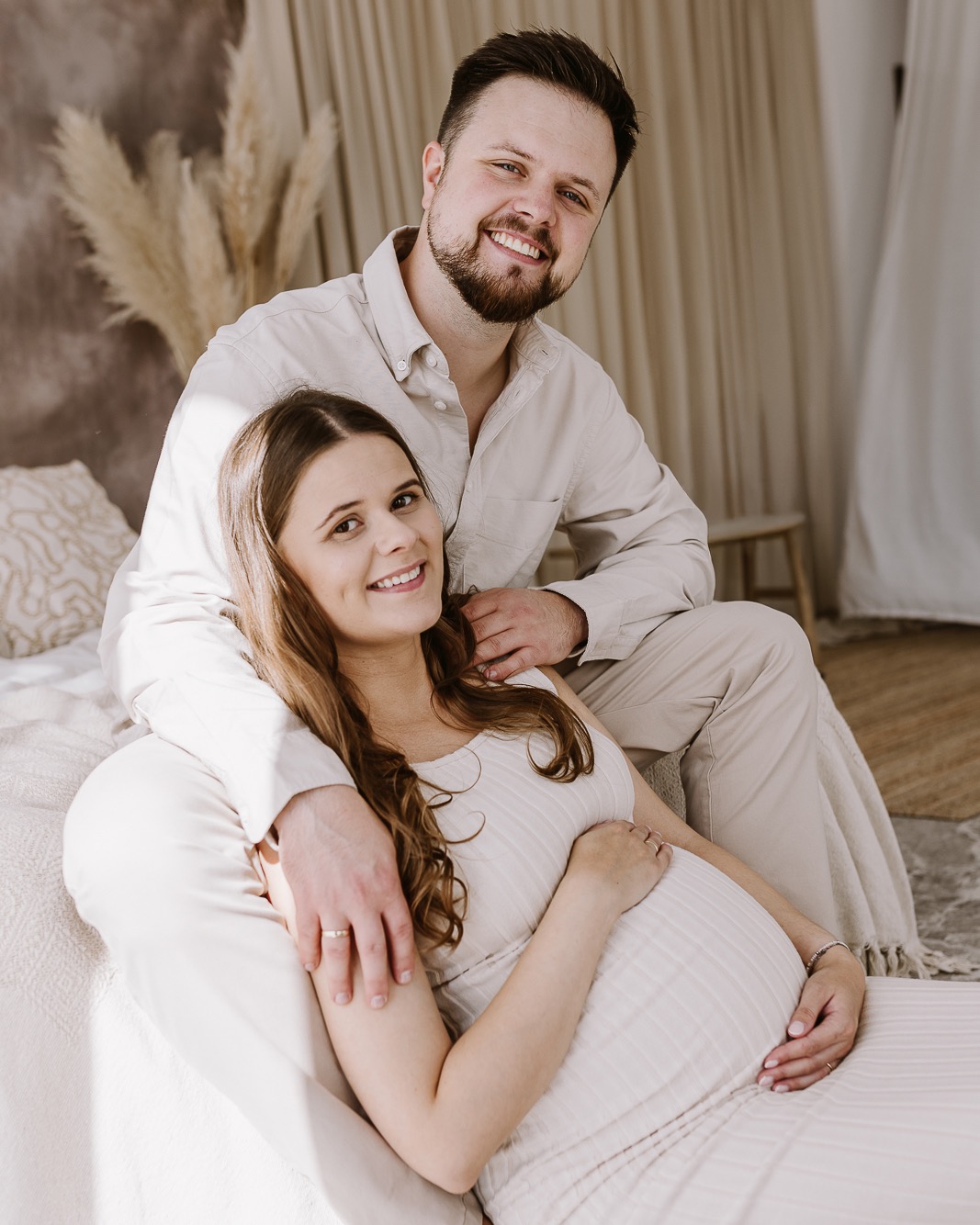 Uśmiechnięta para w jasnych ubraniach: kobieta w ciąży siedzi, mężczyzna obejmuje ją ramieniem. Naturalne światło, beżowe tło, pampasowa trawa w wazonie.