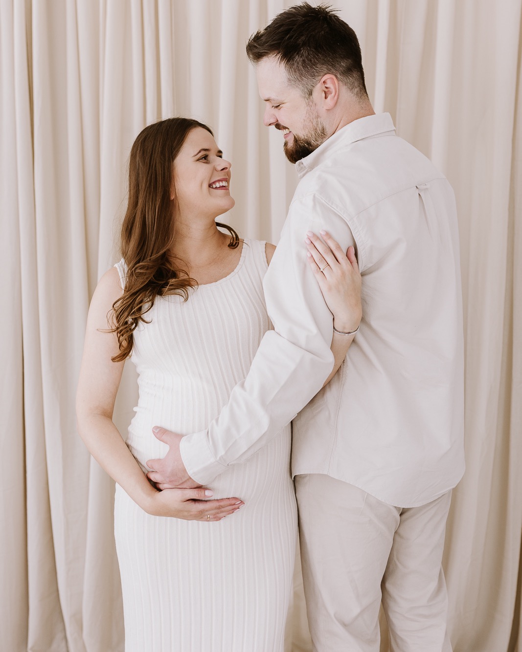 Uśmiechnięta para w jasnych ubraniach, kobieta w ciąży trzymająca dłoń na brzuchu, na tle kremowych zasłon. Delikatne światło, ciepła atmosfera.