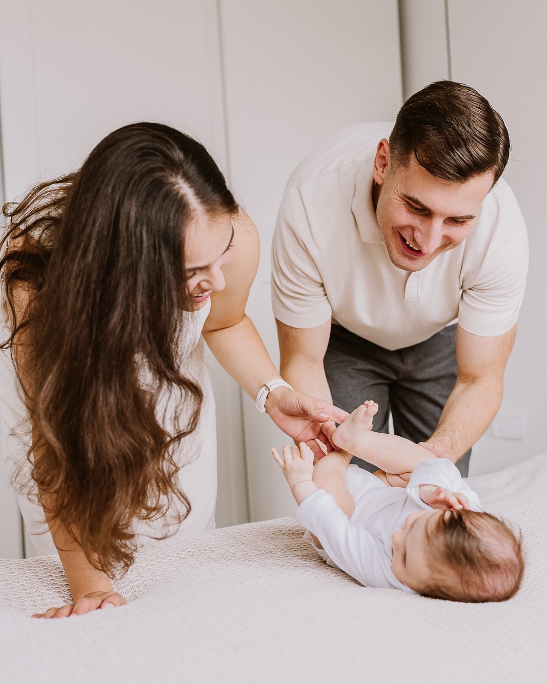 Uśmiechnięci rodzice bawią się z leżącym na plecach niemowlęciem w białym body na beżowym kocu. Jasne, minimalistyczne wnętrze z białymi drzwiami w tle.