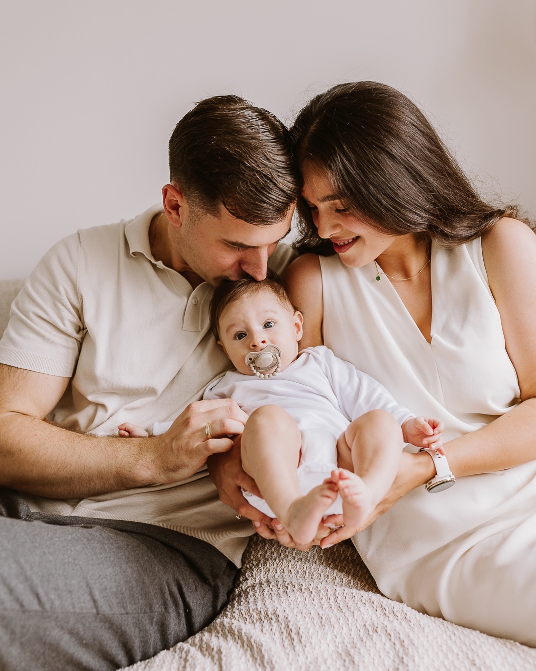 Rodzinne ujęcie: rodzice w jasnych ubraniach czule obejmują niemowlę z smoczkiem, siedząc na beżowej kanapie. Tata całuje dziecko w czoło, mama patrzy z uśmiechem.