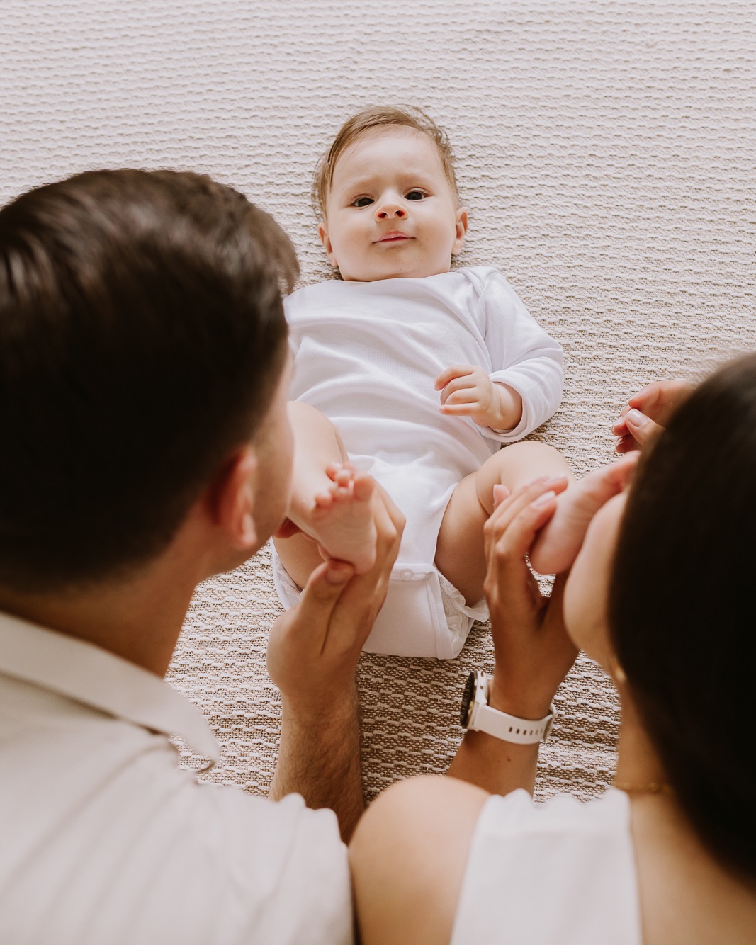 Rodzinne ujęcie z góry: rodzice całują stopy leżącego na beżowym tle niemowlęcia w białym body. Mama ma biały zegarek, tata białą koszulę.