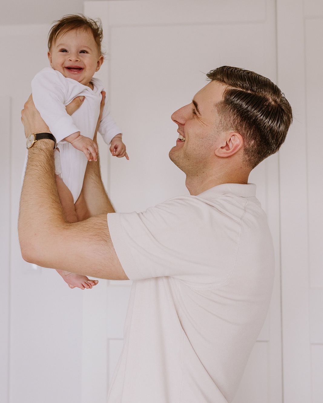 Uśmiechnięty tata podnosi w górę roześmiane dziecko w białym body na tle białych drzwi. Mężczyzna ma na sobie beżową koszulkę polo i zegarek.