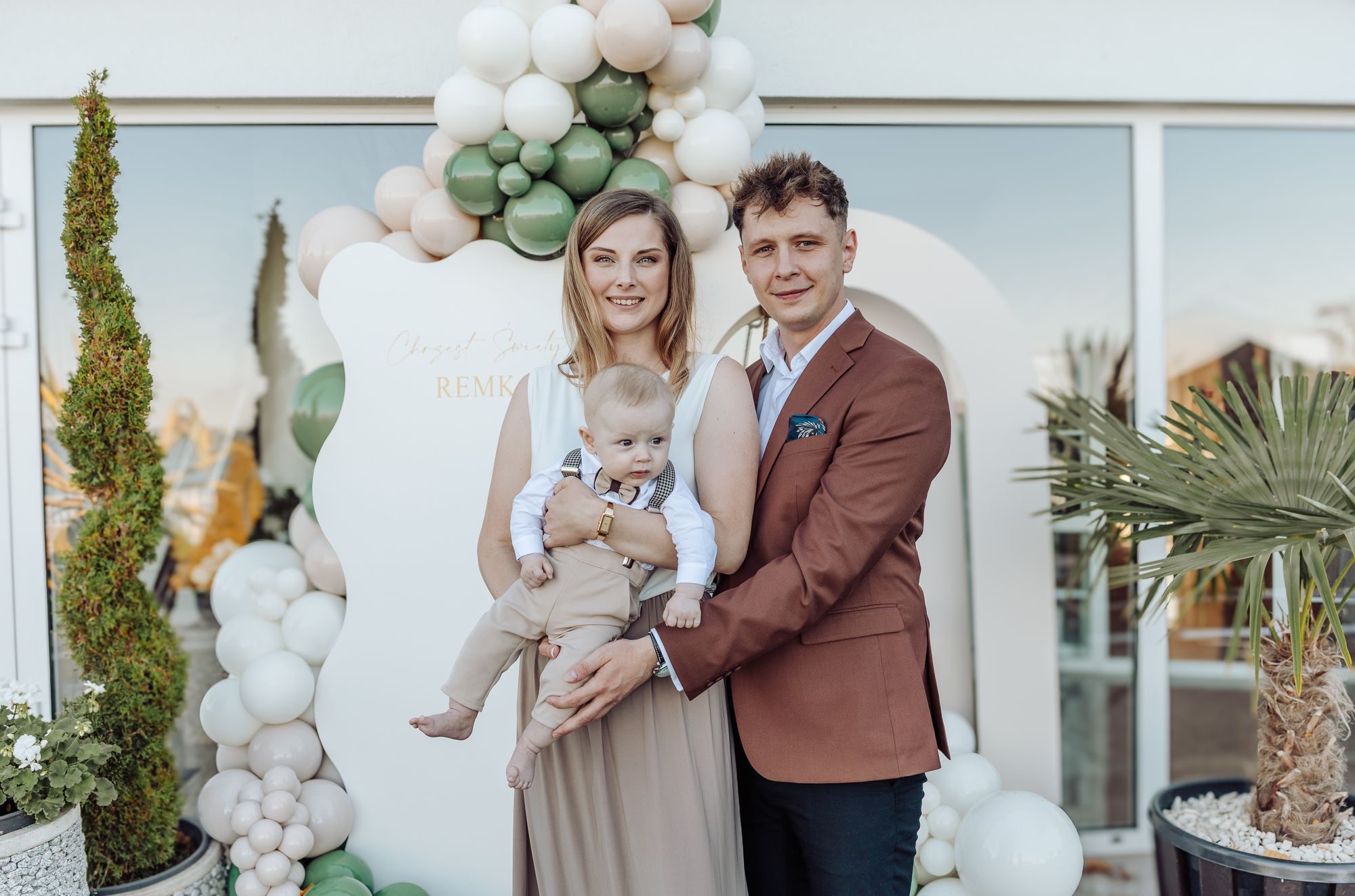 Uśmiechnięta rodzina z małym dzieckiem pozuje na tle dekoracji z balonów w pastelowych kolorach. Ojciec w brązowej marynarce, matka w beżowej sukience, dziecko w eleganckim ubranku.