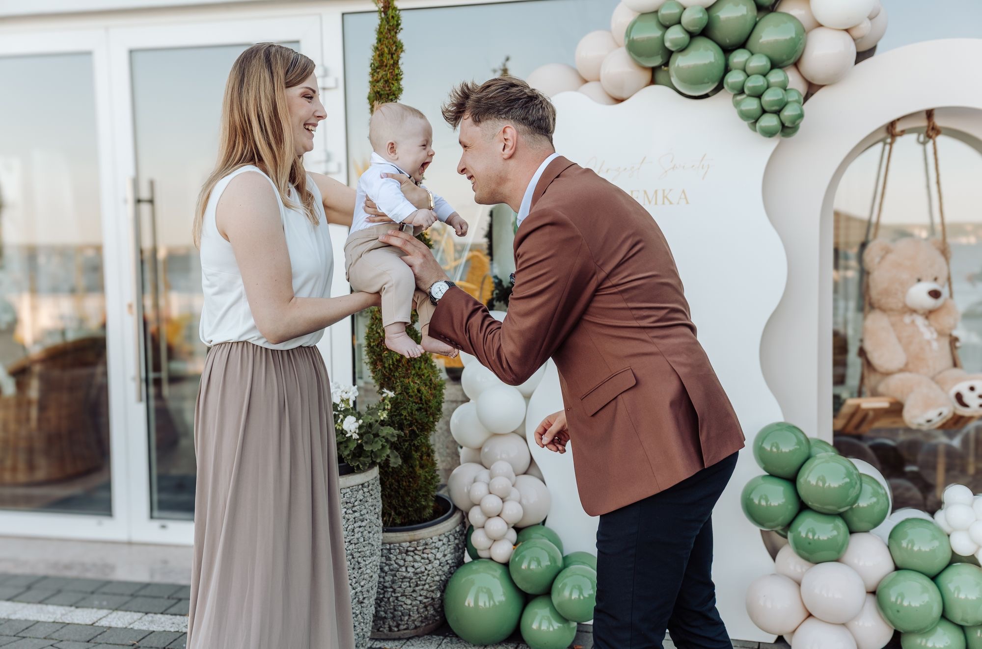 Uśmiechnięta rodzina z niemowlęciem na tle dekoracji balonowych i ścianki z napisem. Sesja rodzinna w plenerze, jasne kolory, radosna atmosfera.