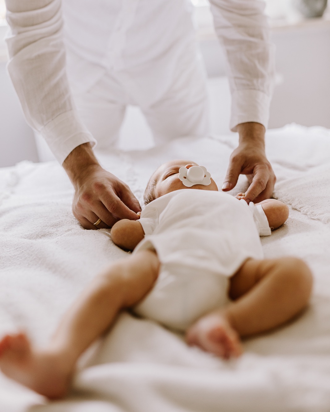 Noworodek leży na białym kocyku z smoczkiem w ustach, dłonie rodzica delikatnie go dotykają. Ujęcie z góry, miękkie światło, dominująca biel.