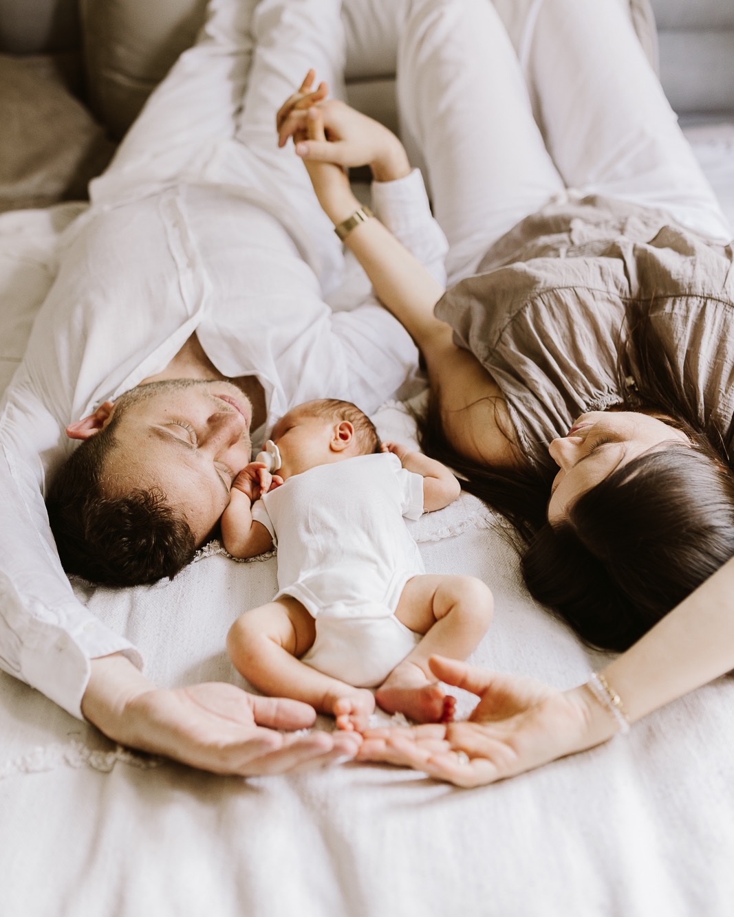 Rodzinna fotografia noworodkowa: rodzice leżący obok śpiącego dziecka na białym prześcieradle, delikatne światło, ciepła kolorystyka, widok z góry.