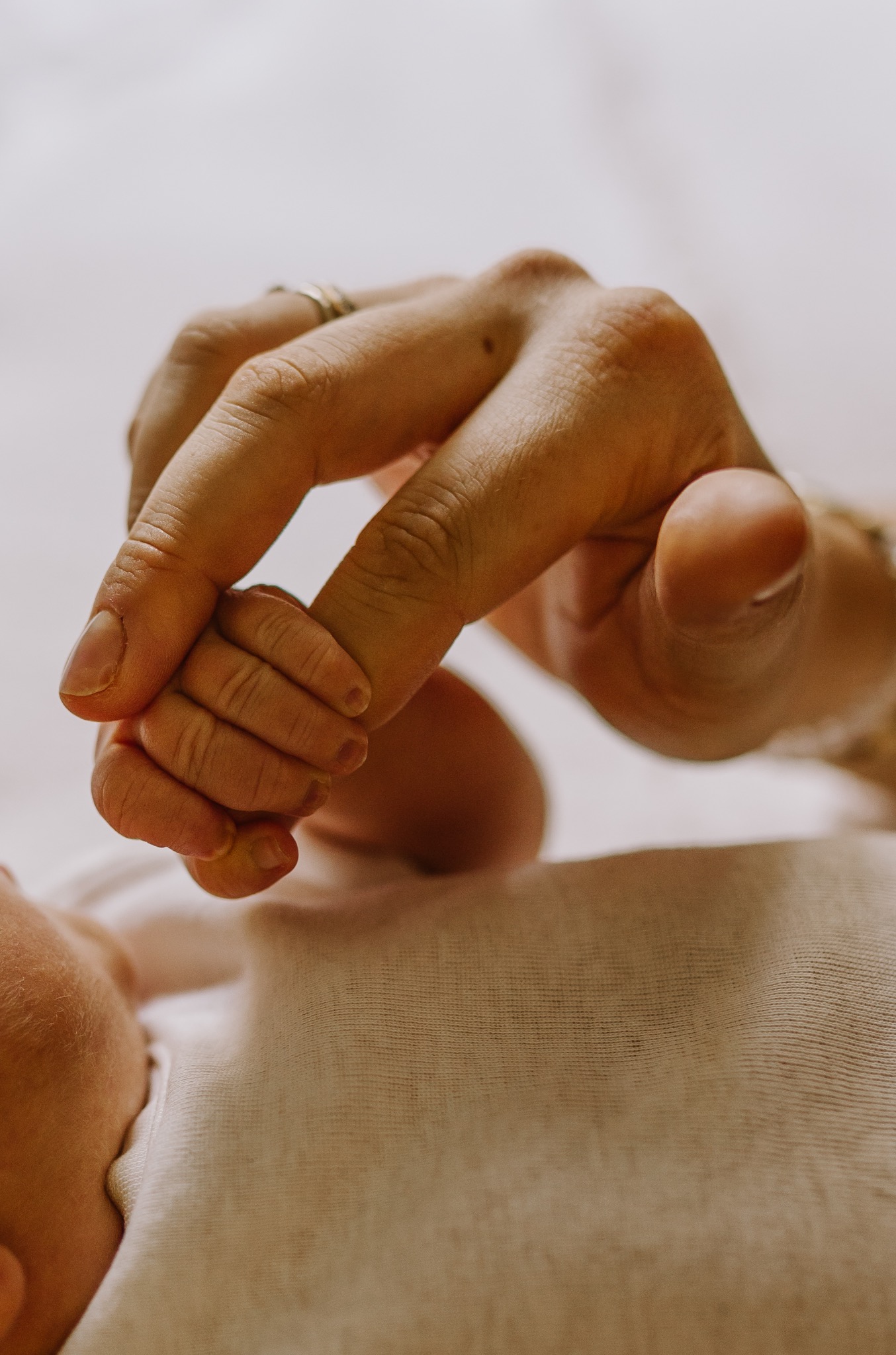 Dłoń matki z obrączką delikatnie trzyma drobną rączkę noworodka na kremowym tle. Bliski kadr podkreśla więź i czułość momentu.