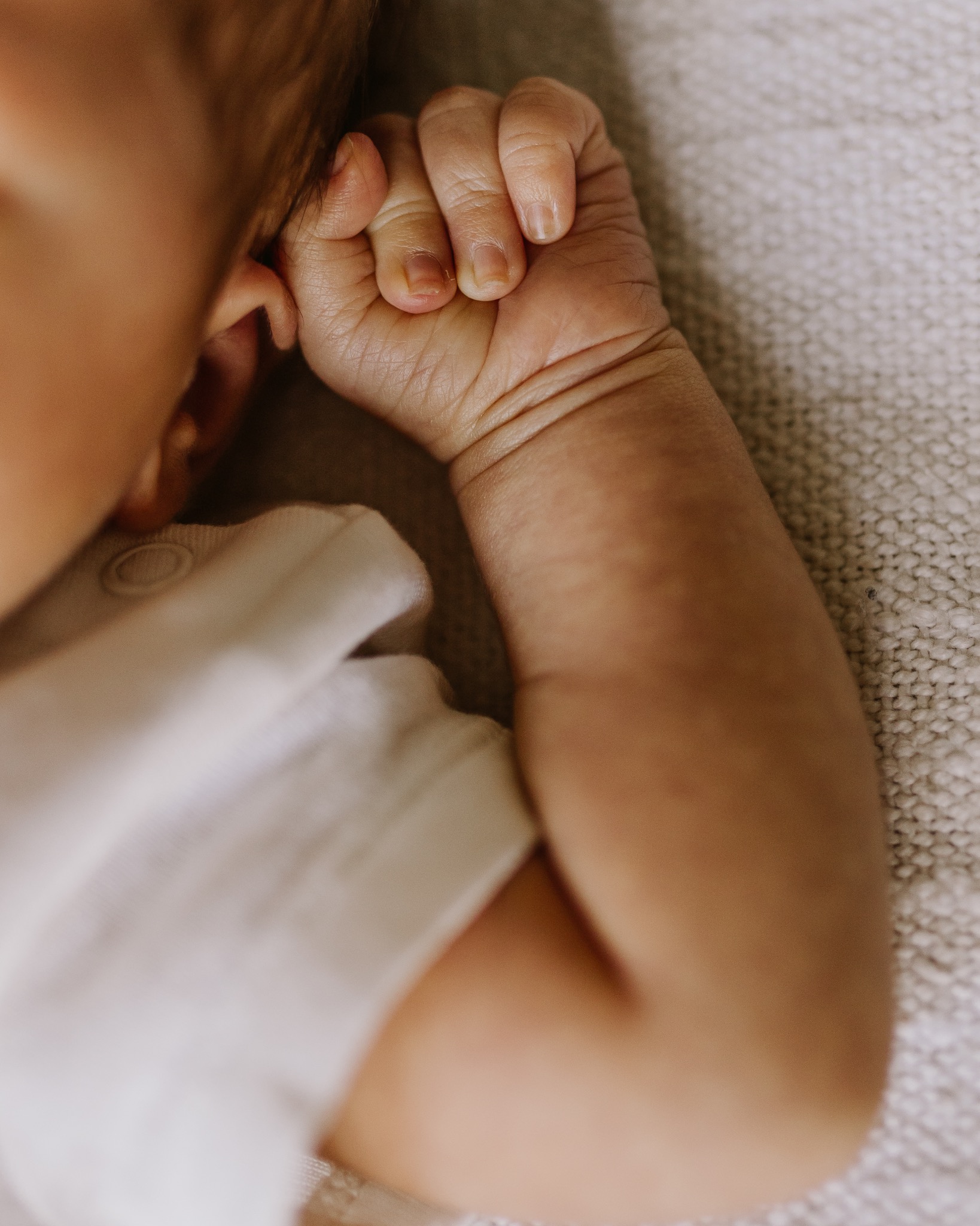 Dłoń noworodka zaciśnięta w piąstkę, leżąca na beżowym, dzierganym kocyku. Widoczny fragment twarzy i ubranka dziecka. Delikatne, naturalne światło.