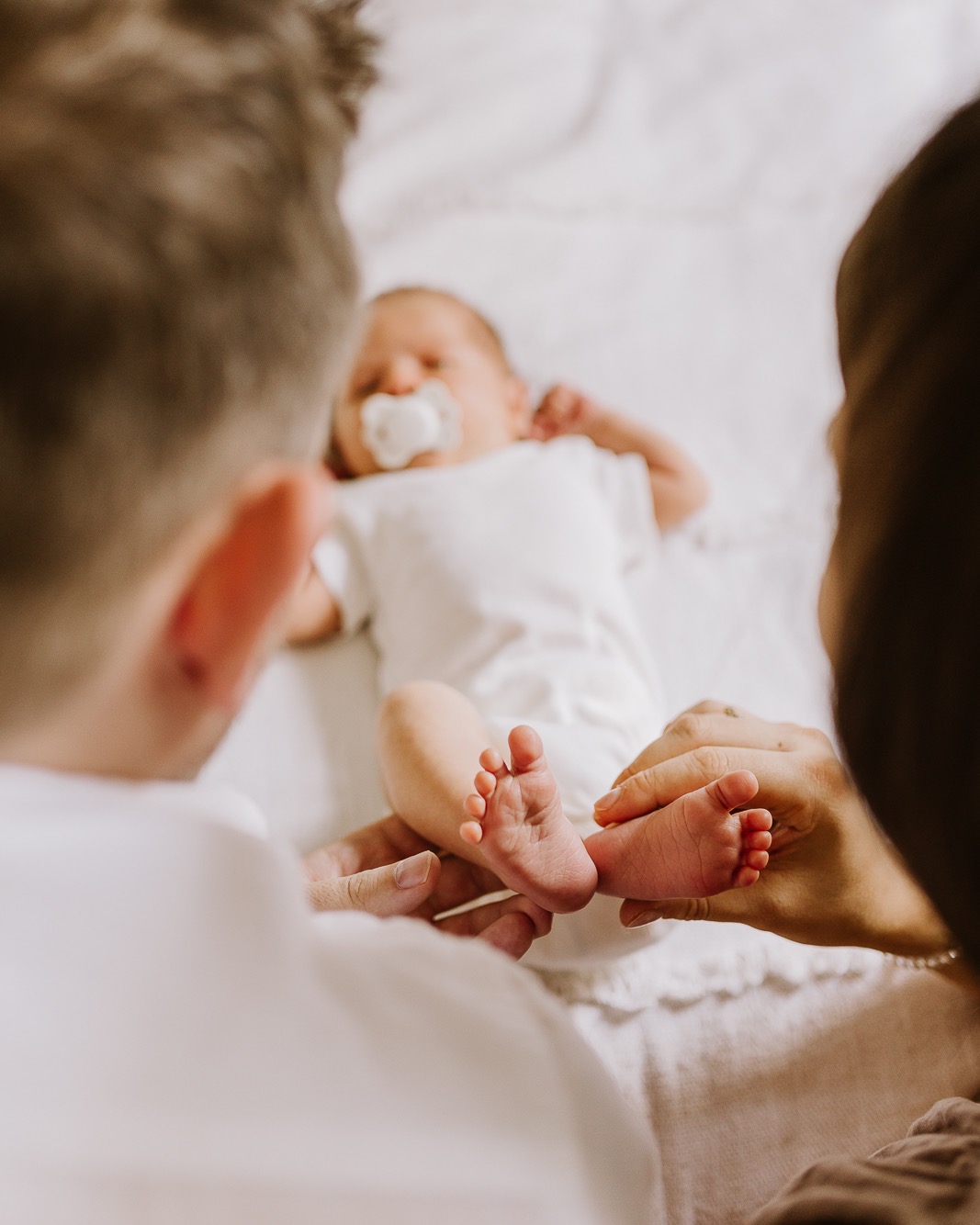 Rodzice trzymają stopy noworodka leżącego na białym kocyku. Ujęcie zza pleców rodziców, skupione na małych stopach dziecka i delikatnych dłoniach.