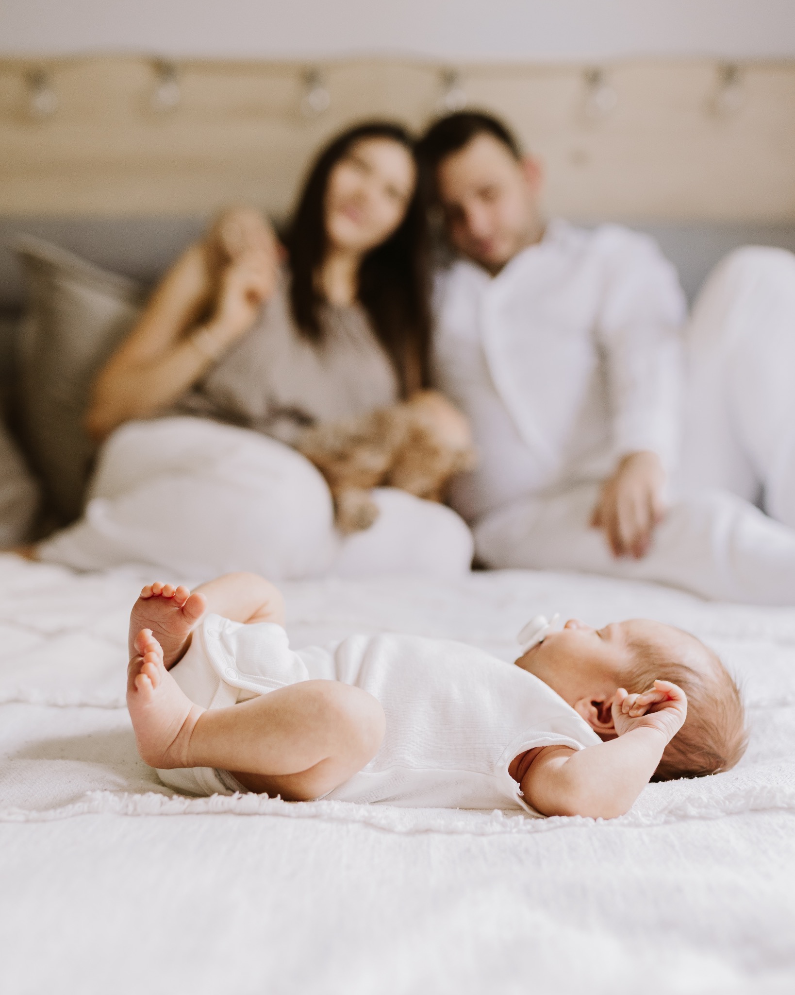 Noworodek w białym ubranku leży na łóżku, w tle rozmyta sylwetka rodziców. Delikatne, naturalne światło podkreśla intymność rodzinnej sesji.