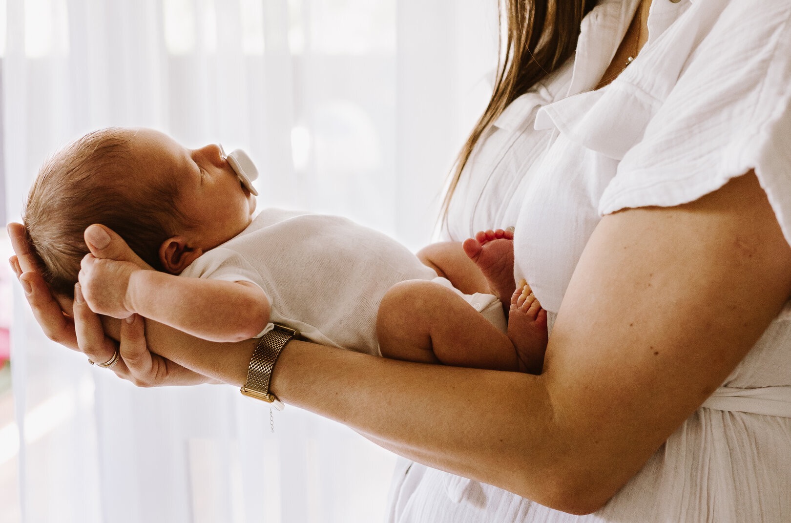 Delikatne ujęcie noworodka z smoczkiem w ramionach mamy, w jasnym wnętrzu w Warszawie. Dziecko ubrane w białe body, subtelne światło podkreśla detale.