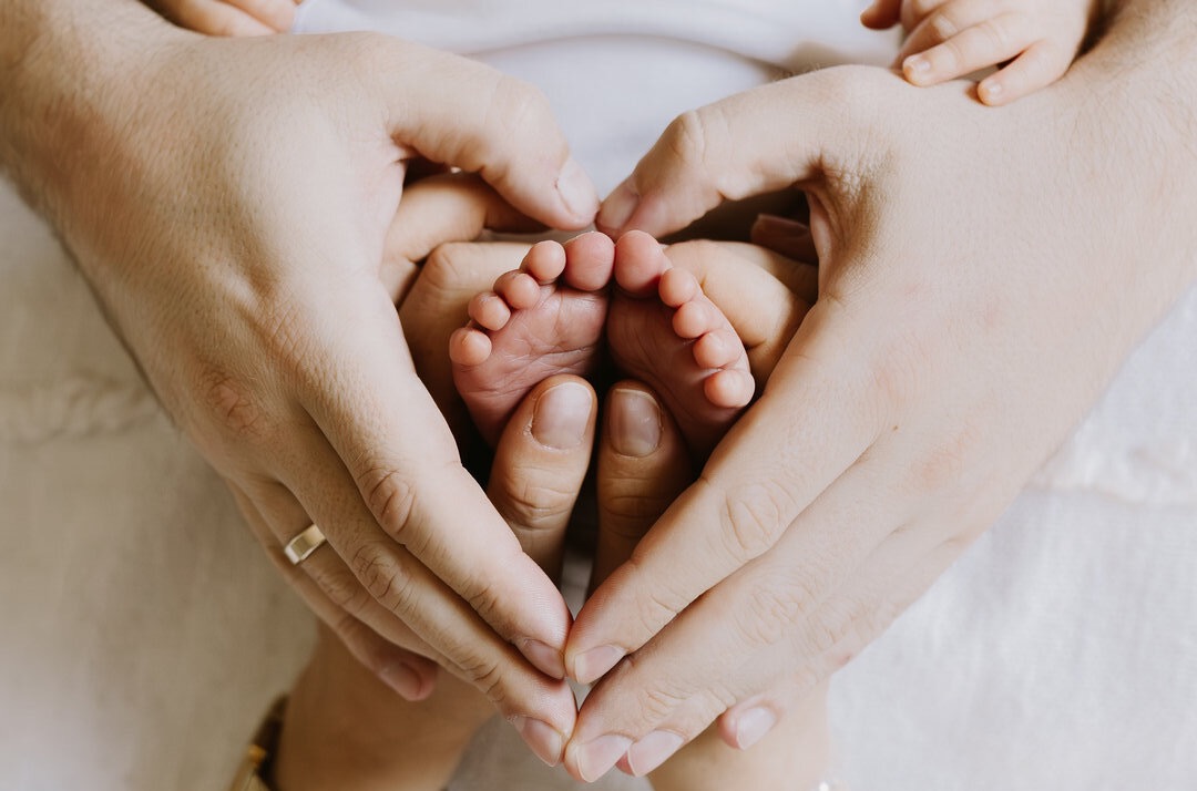 Dłonie rodziców formują serce wokół stópek noworodka, delikatne światło podkreśla detale skóry, symbolizując miłość i więź rodzinną. Ujęcie z bliska.