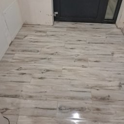 Płytkarz Andrii Kramarenko - Ułożona podłoga z płytek imitujących drewno, widok z góry, przy ścianie i drzwiach. Jasne płytki z ciemniejszymi smugami, błyszczące wykończenie.