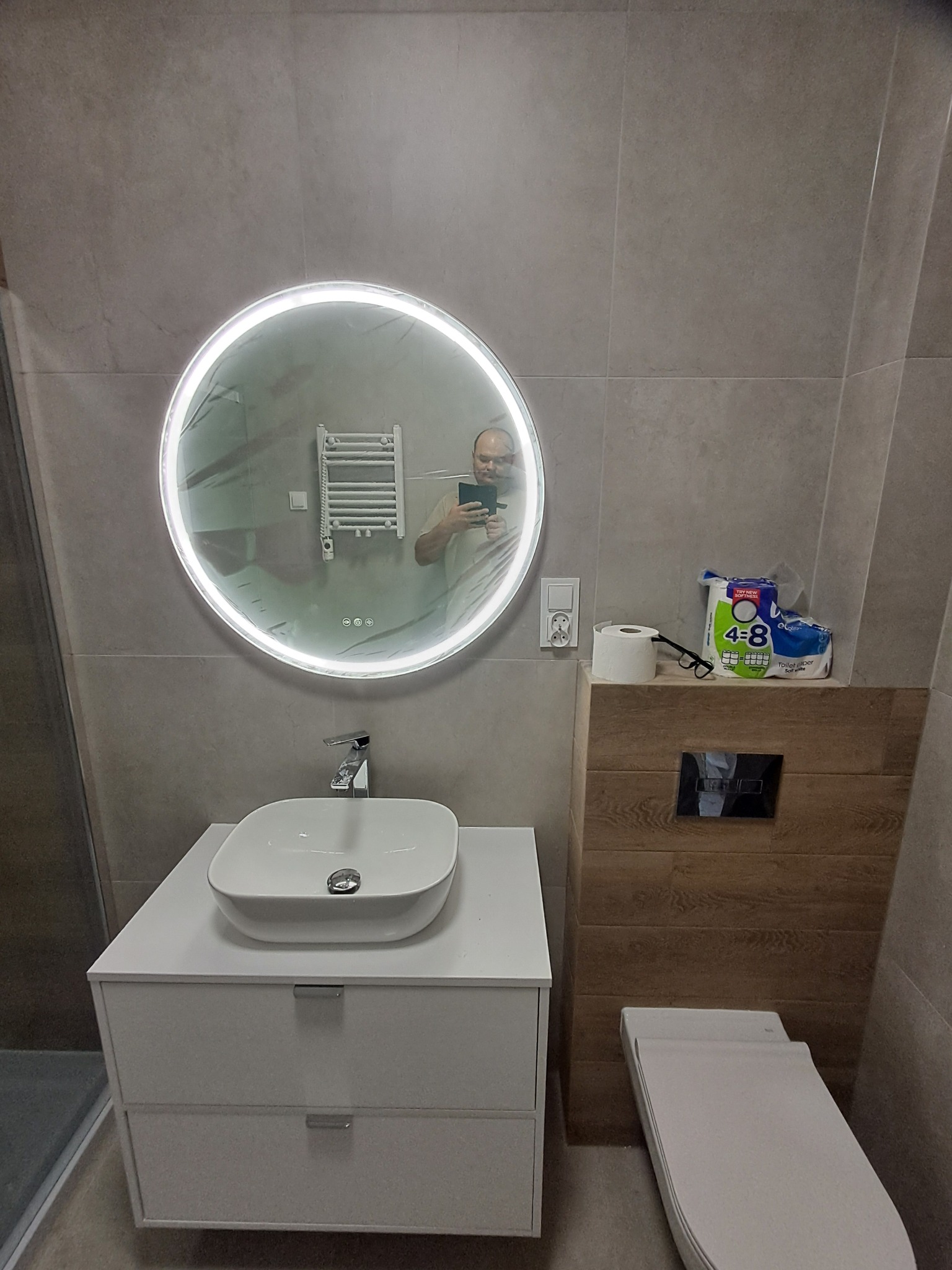 Nowoczesna łazienka z umywalką nablatową na białej szafce, okrągłym lustrem z podświetleniem LED i podwieszaną toaletą. Szare płytki na ścianach.