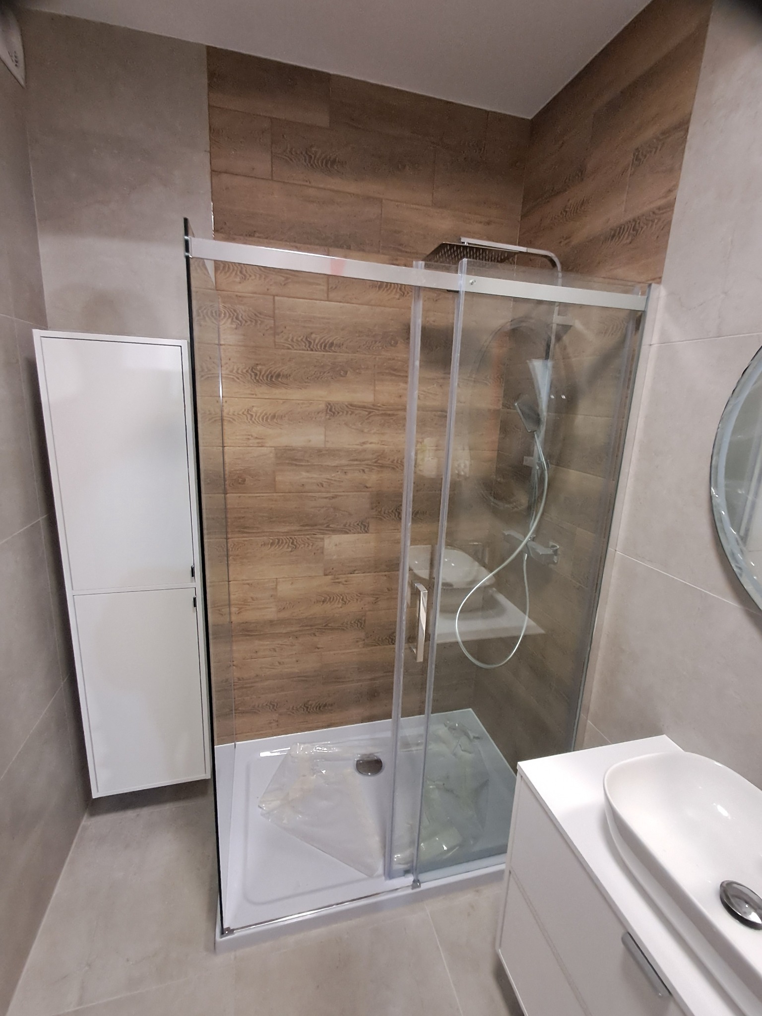 Nowoczesna łazienka po remoncie: kabina prysznicowa ze szklanymi drzwiami, biała szafka z umywalką i wysoka szafka. Płytki imitujące drewno na ścianie.