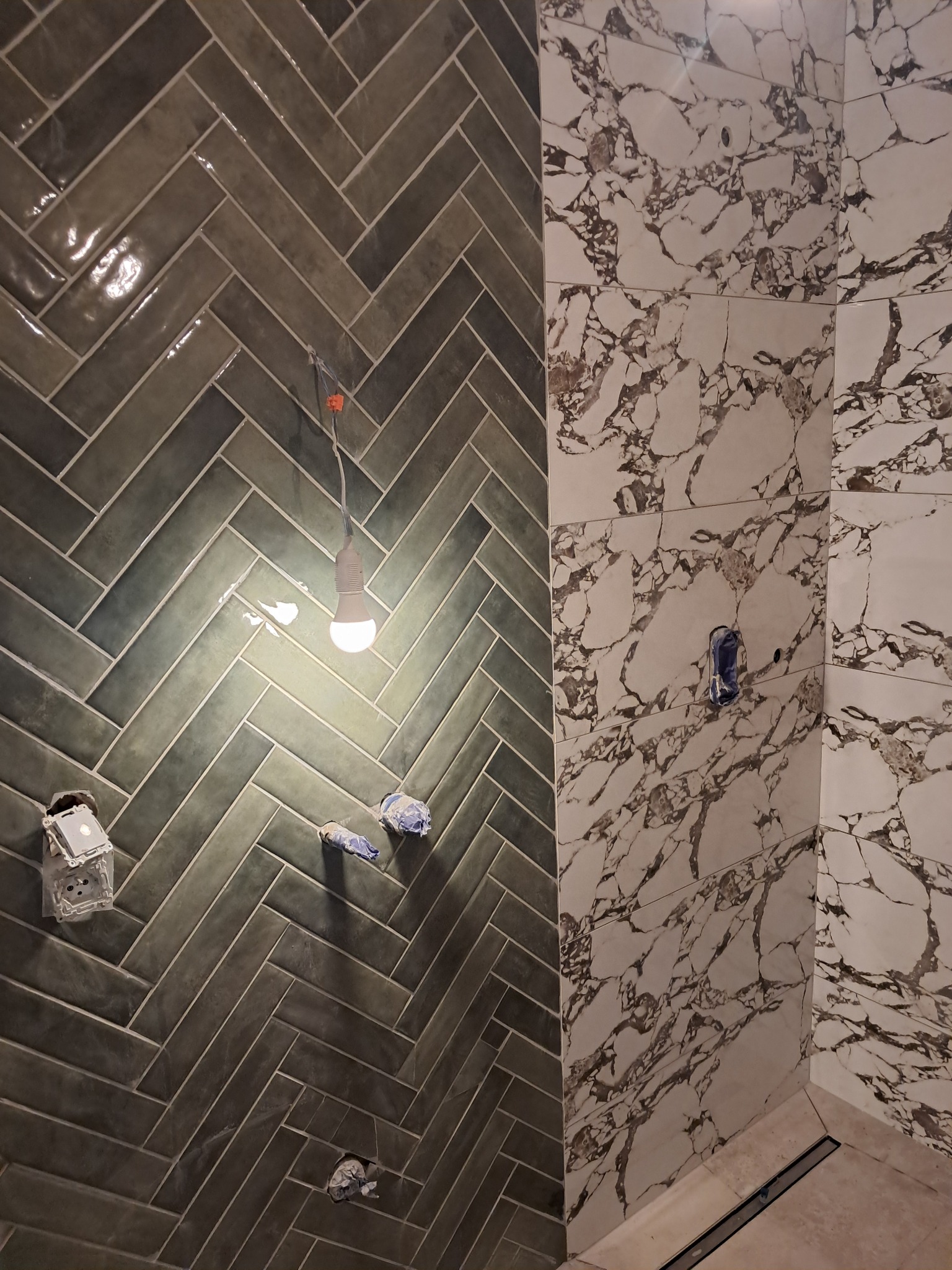 Ściana łazienki wyłożona płytkami w dwóch wzorach: jodełka w odcieniach szarości i imitacja marmuru. Widoczne puszki elektryczne i tymczasowe oświetlenie.