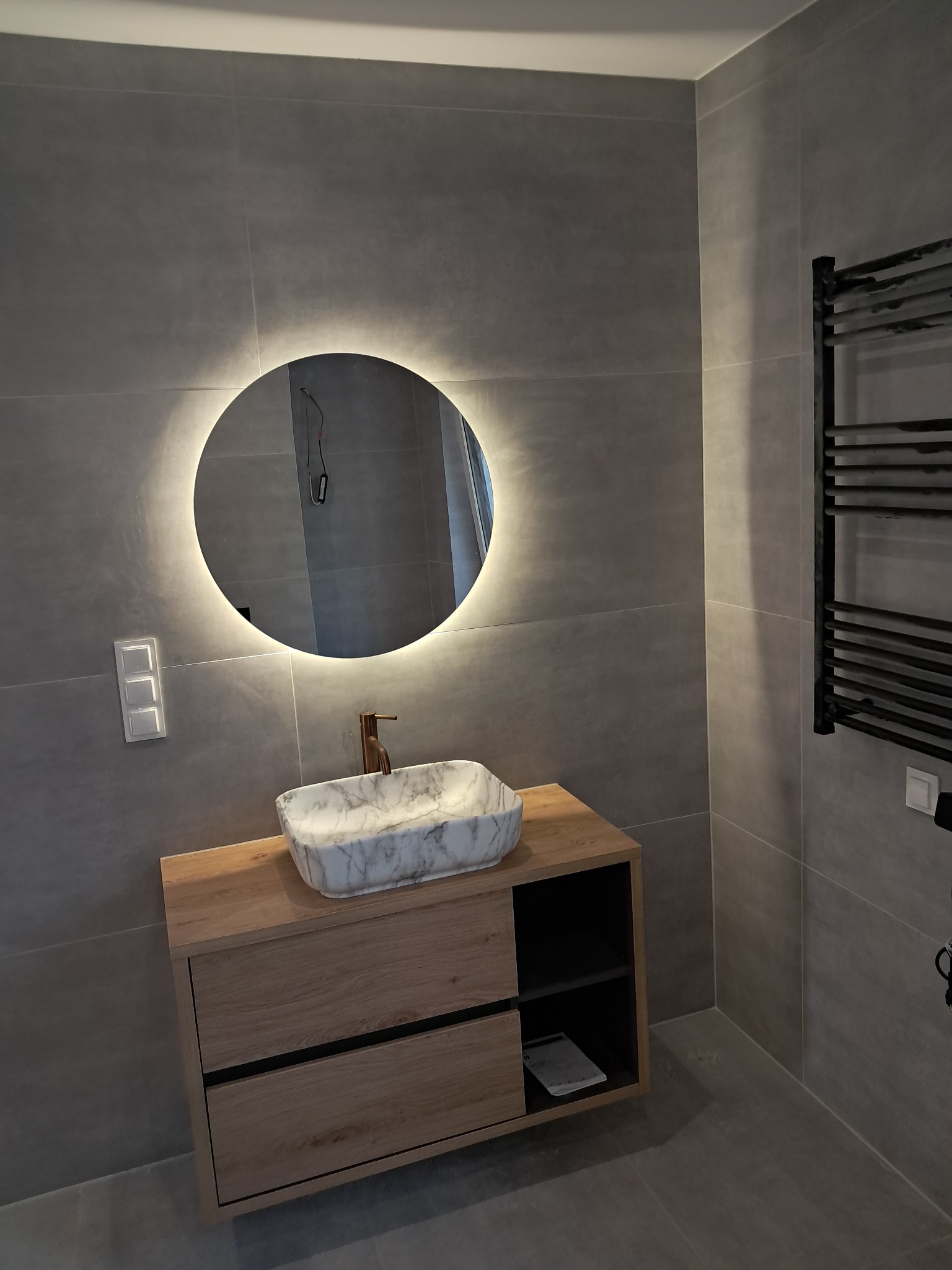 Nowoczesna łazienka z szarą płytką, okrągłym lustrem z podświetleniem LED, marmurową umywalką na drewnianej szafce i czarnym grzejnikiem drabinkowym.