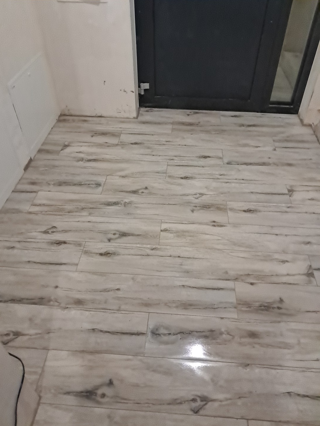 Ułożona podłoga z płytek imitujących drewno, widok z góry, przy ścianie i drzwiach. Jasne płytki z ciemniejszymi smugami, błyszczące wykończenie.