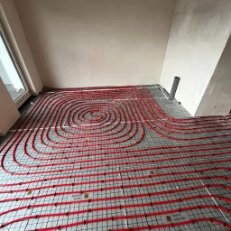 Usługi Hydrauliczne Konrad Bukowski - Czerwone rury ogrzewania podłogowego ułożone spiralnie na szarej macie z czarną siatką w surowym wnętrzu. Widok z góry, instalacja przed wylaniem posadzki.