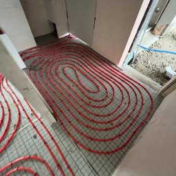 Usługi Hydrauliczne Konrad Bukowski - Czerwone rury ogrzewania podłogowego ułożone na siatce w surowym wnętrzu, widok z góry, etap instalacji przed wylewką.