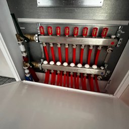 Usługi Hydrauliczne Konrad Bukowski - Rozdzielacz ogrzewania podłogowego z czerwonymi rurkami, zamontowany w szafce. Widoczne zawory, odpowietrzniki i elementy mocujące. Ujęcie z bliska, z otwartą szafką.
