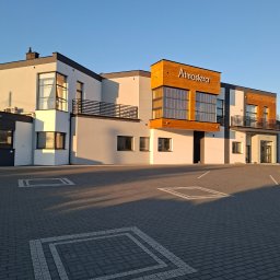 MMAM - Nowoczesny budynek z elementami drewnianymi i balkonami, oświetlony popołudniowym słońcem. Widoczny napis 'Atmosfera' na elewacji i kostka brukowa na parkingu.