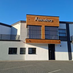 MMAM - Nowoczesny budynek z białą elewacją, drewnianym akcentem i napisem 'Atmosfera' pod błękitnym niebem. Widoczny fragment parkingu z kostki brukowej.