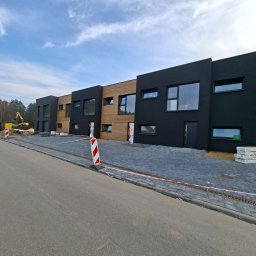 Mmam - Nowoczesny szeregowiec z czarną i drewnianą elewacją, kostka brukowa, widoczne prace ziemne w tle. Minimalistyczny design, słoneczny dzień.