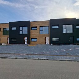 Mmam - Nowoczesna elewacja szeregowca z czarnymi i drewnianymi panelami, duże okna, kostka brukowa na podjeździe, zabezpieczone drzwi wejściowe.