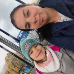 Klaudia Klaudia - Uśmiechnięta kobieta i dziewczynka w czapce z kotem, patrzące przez okno z widokiem na jesienne drzewa. Nietypowy kąt ujęcia, naturalne światło.