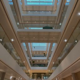 Detech - Wnętrze nowoczesnego biurowca z atrium, widok z dołu na szklany dach i liczne kondygnacje. Białe ściany, szklane balustrady, schody i rośliny doniczkowe.