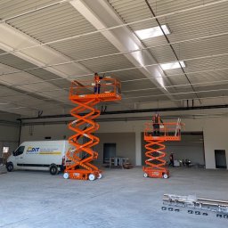 Detech - Montaż instalacji w hali: Dwóch pracowników na podnośnikach nożycowych, montuje elementy pod sufitem. Biała hala z widocznymi instalacjami i oświetleniem.