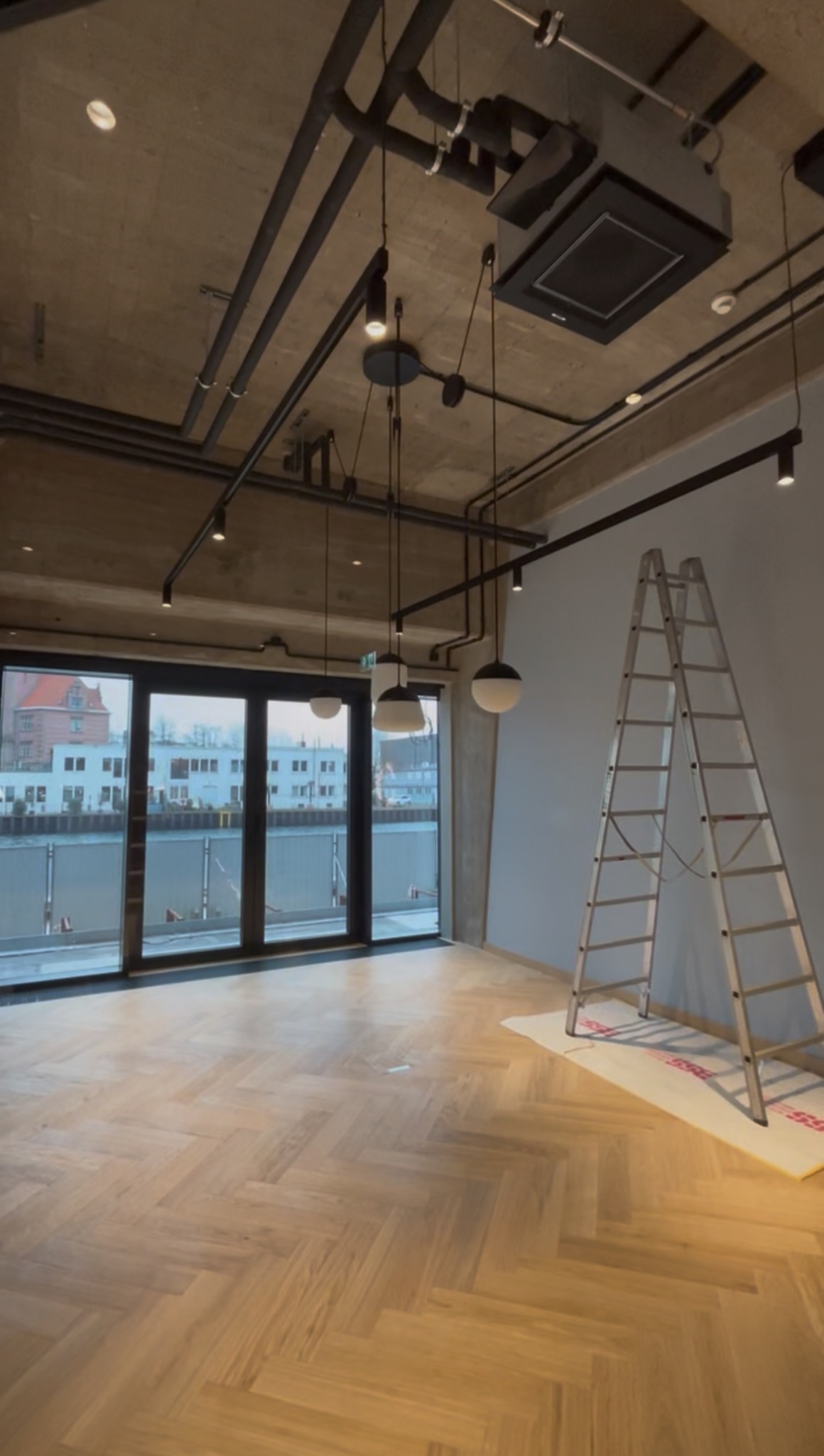 Wnętrze z parkietem w jodełkę, surowym betonowym sufitem z instalacjami i nowoczesnym oświetleniem. Aluminiowa drabina oparta o ścianę. Widok na rzekę przez okna.